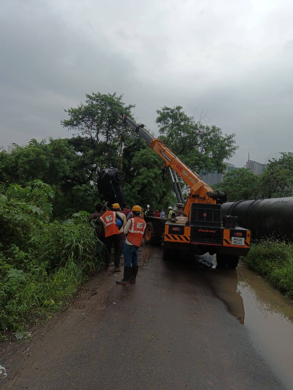A black overturned car being lifted from a ditch or embankment by a large orange mobile crane, with rescue workers in safety vests on a muddy road.