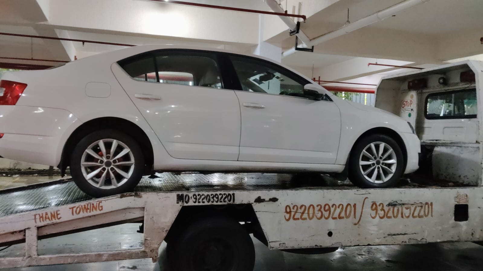 White sedan loaded onto a tow truck inside a covered parking structure, highlighting the flatbed's contact numbers.
