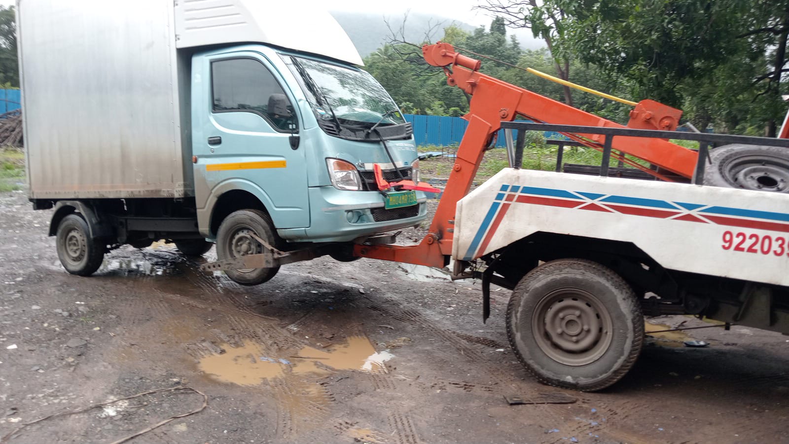 Small commercial mini-truck being towed by a wheel-lift truck on a muddy, unpaved commercial road.