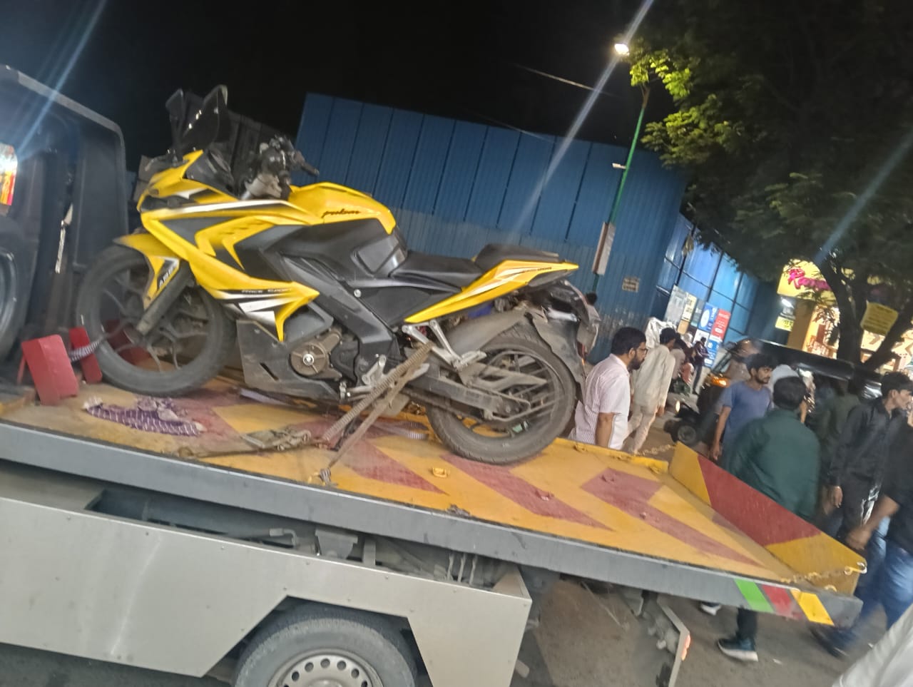 Bright yellow Bajaj Pulsar RS200 sport bike secured on a flatbed tow truck at night in a crowded street.