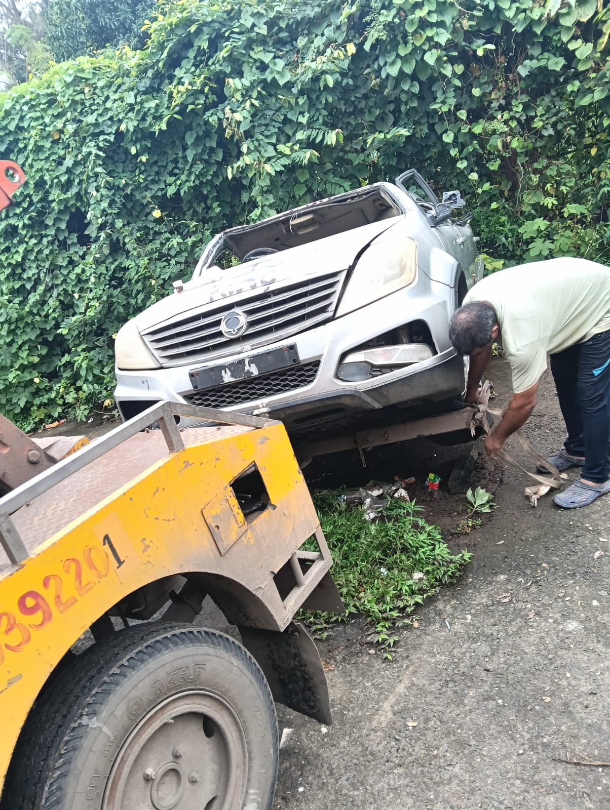 Siddhivinayak Towing technician recovering a heavily damaged vehicle near a leafy roadside area.