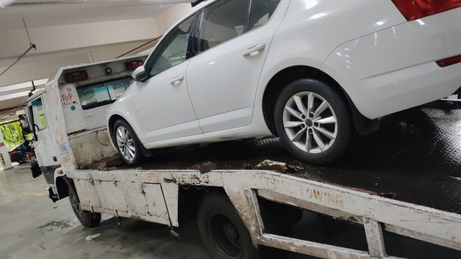 White Kia Seltos SUV on a wet flatbed tow truck ramp, parked near a residential building in the rain.